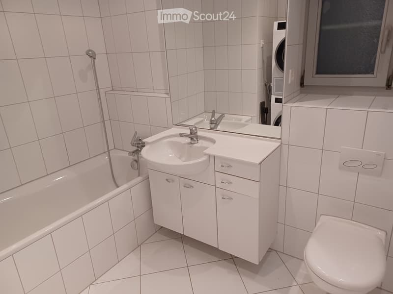 Modern bathroom with white tiles, a bathtub, a sink with a cabinet, a toilet, and a washing machine in the corner.