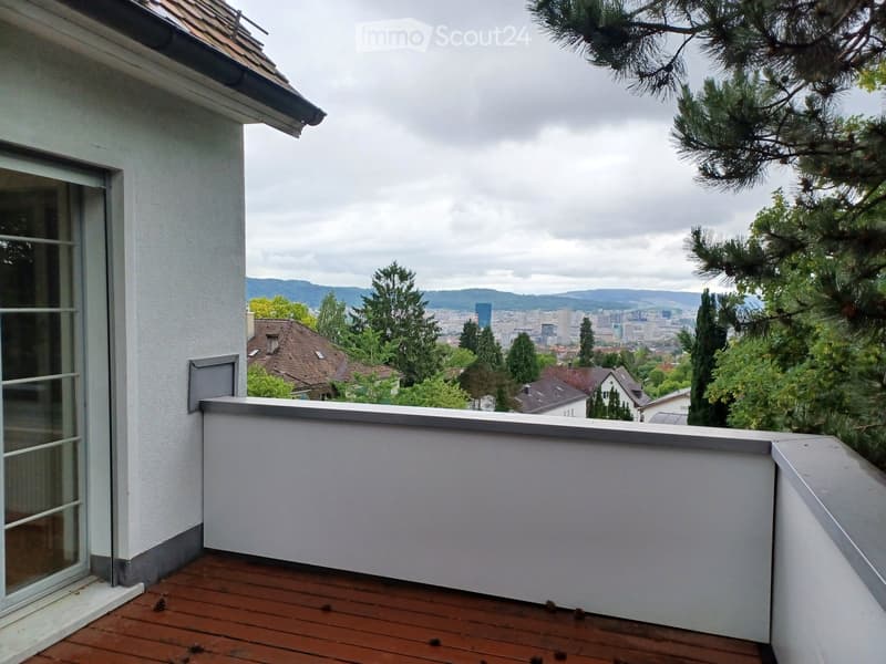 Balcony with wooden decking, white railing, and a view of a cityscape with trees and hills in the background.