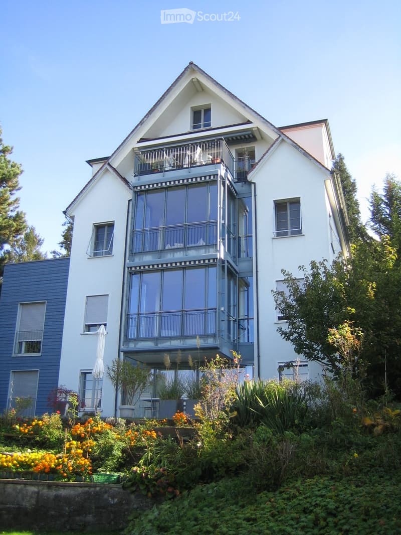 Two-story house with white exterior, large balcony, and garden area with colorful flowers and greenery.