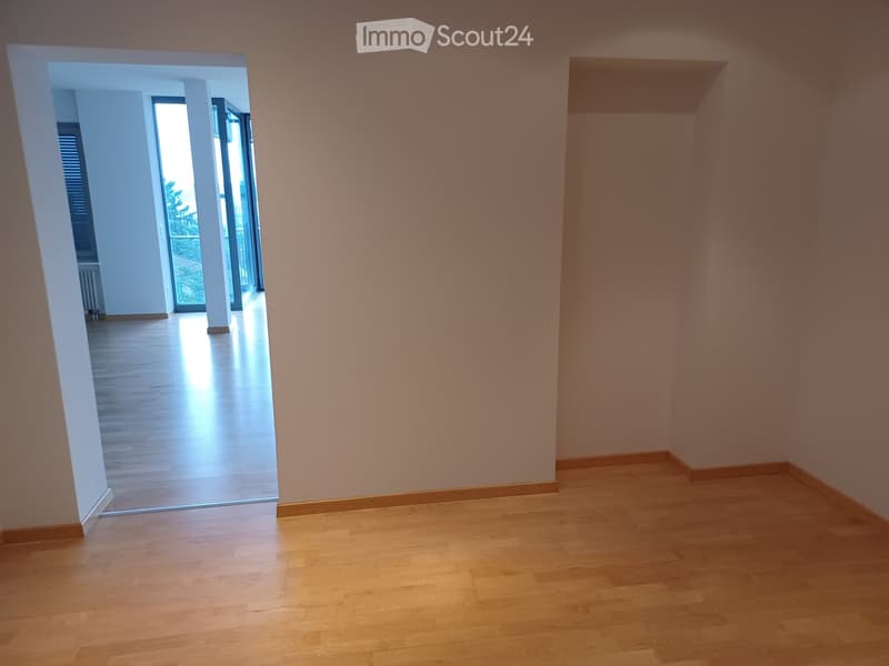 Empty room with light wooden flooring and white walls, featuring an open doorway leading to another space with large windows and a view of greenery.