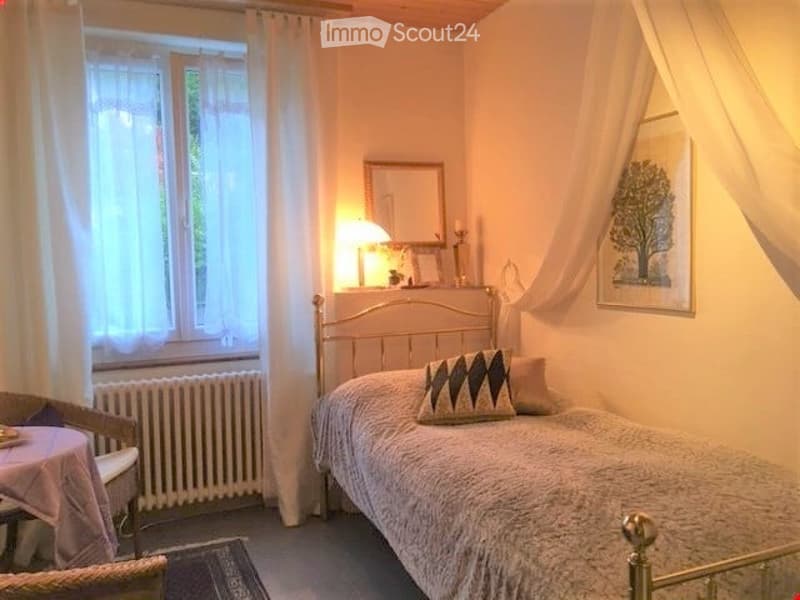 Single bed, lamp, mirror, picture frame, radiator, chair, table, rug, window with curtains