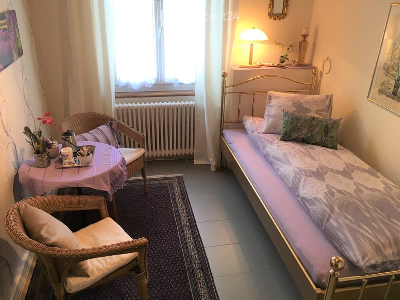 A small bedroom with a single bed, a small table with a lamp, a chair, and a radiator. The room has a carpet, a window with curtains, and a picture frame on the wall.