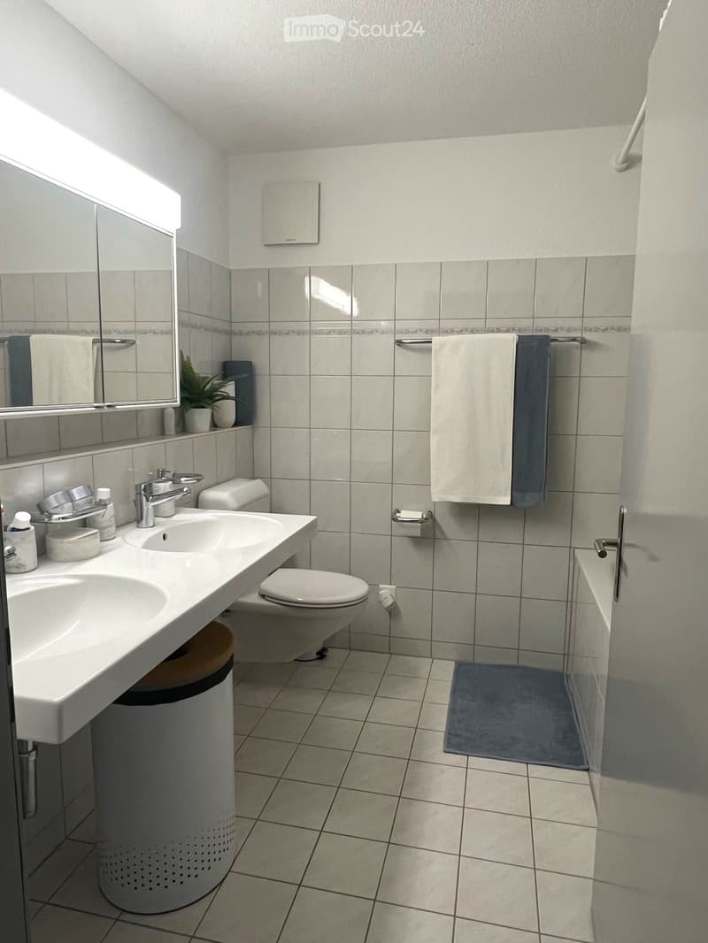 Modern bathroom with a double sink, toilet, bathtub, mirror, and towels. Features tiled walls and floor, a small plant on the windowsill, and a trash can under the sink.