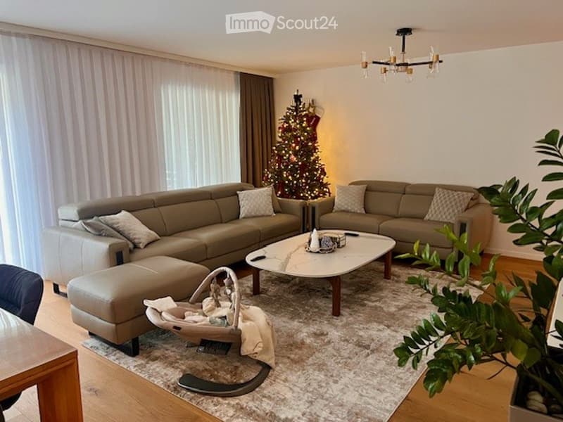 A modern living room with a grey leather sofa, coffee table, baby chair, and a Christmas tree in the corner. The room features large windows with white curtains and a chandelier hanging from the ceiling.