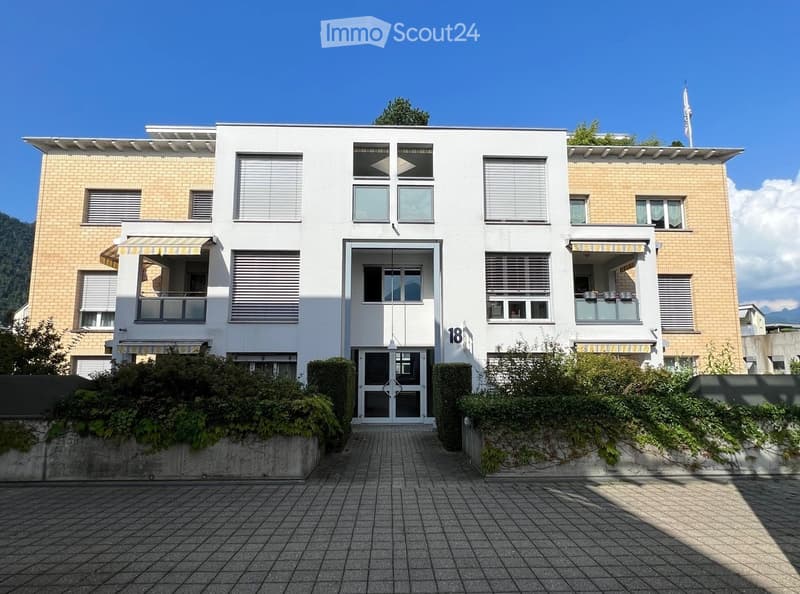 Modern two-story building with white facade, large windows, and balconies. Features a central entrance with glass doors and a small garden area in front.