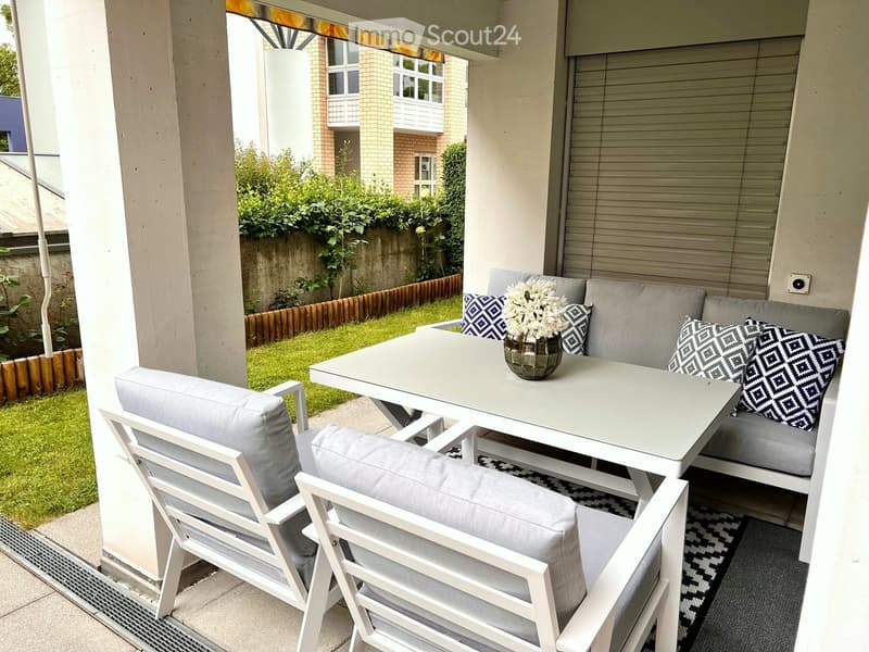Balcony with a white table, two white chairs, a grey sofa, and a vase with white flowers.