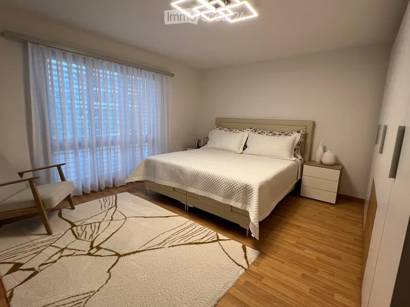 Modern bedroom with a large bed, white bedding, nightstand, wooden chair, and decorative vases. The room features wooden flooring, large windows with sheer curtains, and a white area rug with a leaf design.