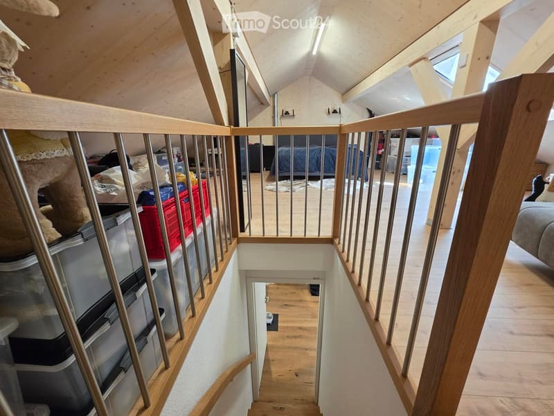 A wooden staircase with metal railings leads up to an attic space. The attic has exposed wooden beams, skylights, and visible storage items including red crates and a bed.