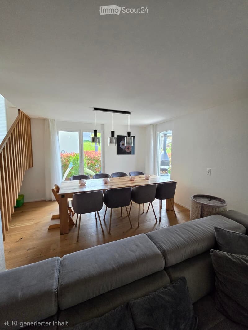 Open-plan living and dining area with a grey sectional sofa, a wooden dining table with six chairs, and a small cat basket. The room features wooden flooring, large windows, and modern pendant lights.