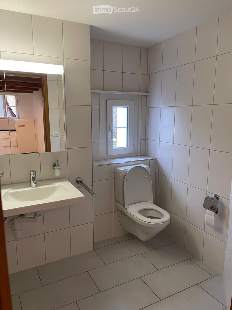 Modern bathroom with white tiled walls and floor, featuring a sink with a chrome faucet, a toilet, a large mirror with built-in lighting, and a small window above the toilet.