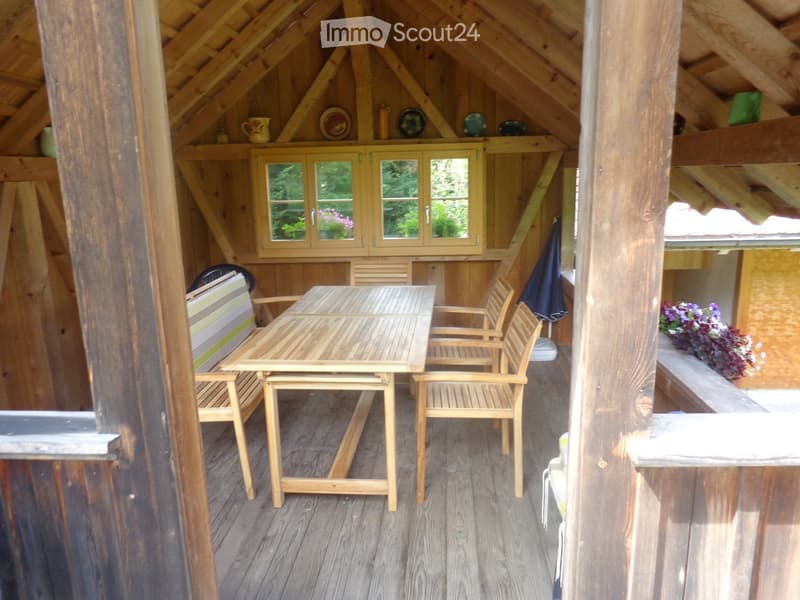 Wooden balcony with a dining table, chairs, and a small cupboard with glass doors. Wooden beams and slanted roof visible.