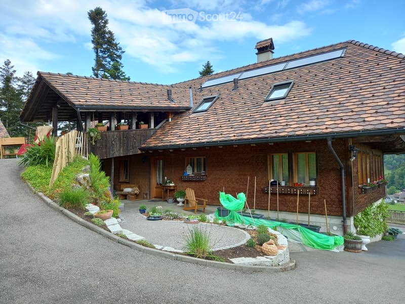 Two-story house with brown tiled roof, multiple windows, and a wooden balcony. The house features a well-maintained garden with potted plants and a wooden chair on the patio.