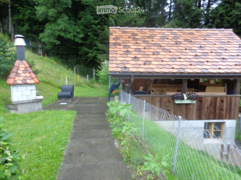 A small wooden outdoor structure with a tiled roof, surrounded by a wire fence, with a barbecue and seating area nearby, set in a green, hilly landscape.