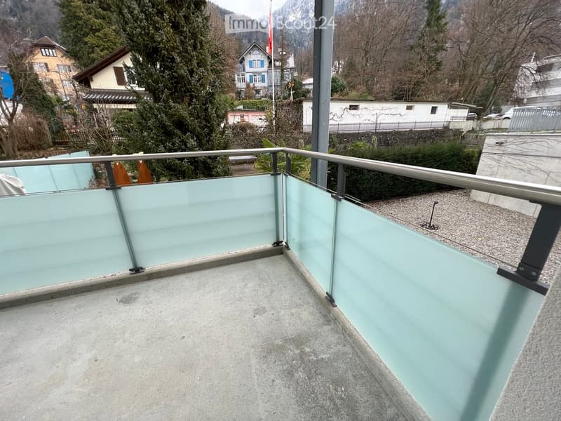 Balcony with glass railing, concrete floor, and a view of surrounding houses and trees.