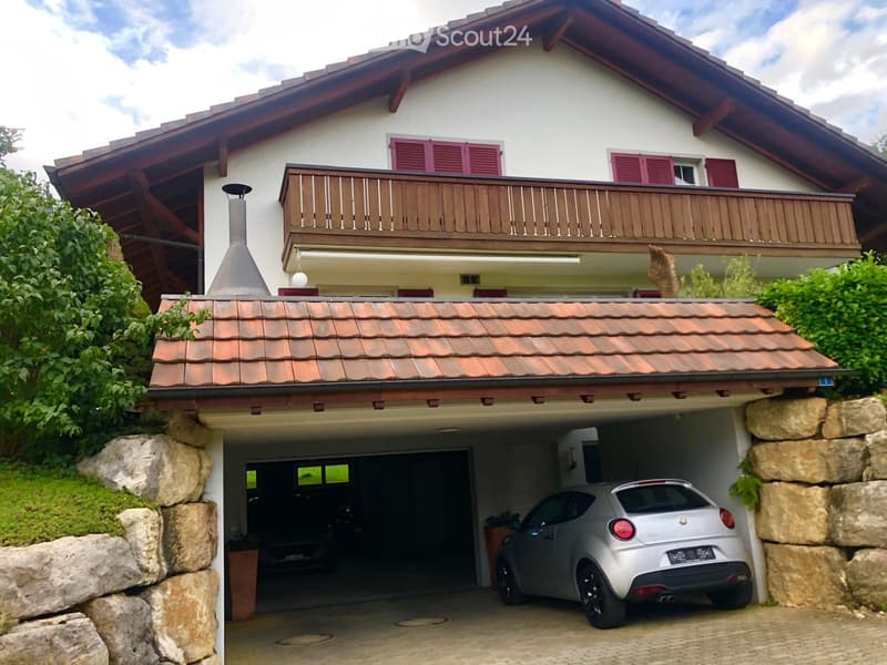 Two-story house with a red tiled roof, wooden balcony, and a garage with a silver car parked inside.