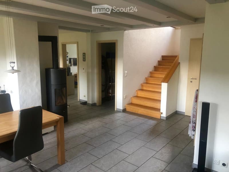 An empty room with a wooden staircase, tiled floor, and a fireplace. There is a table and chair in the left corner.