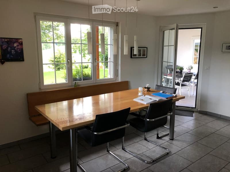 Dining area with a wooden table, chairs, and a bench. There are books, a candle, and picture frames on the table. The room has large windows and an open door leading to a patio.
