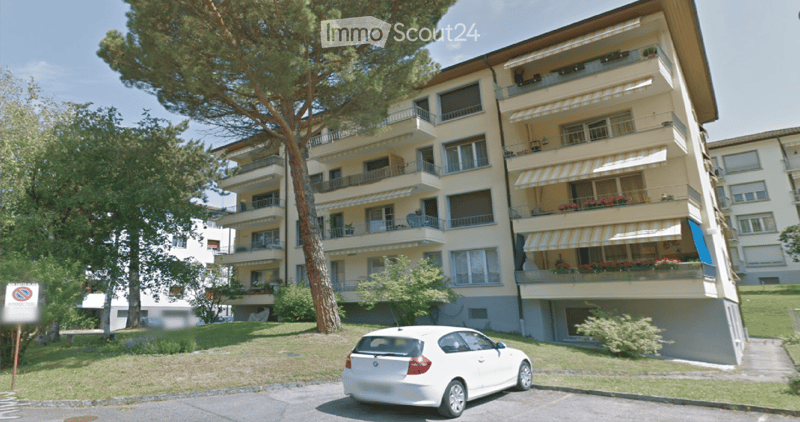 Multi-story residential building with balconies, awnings, and surrounding greenery.