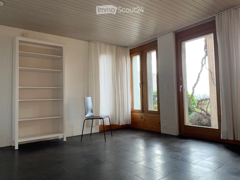Empty room with dark tiled floor, white brick walls, wooden ceiling, large windows with white curtains, and a single modern chair.