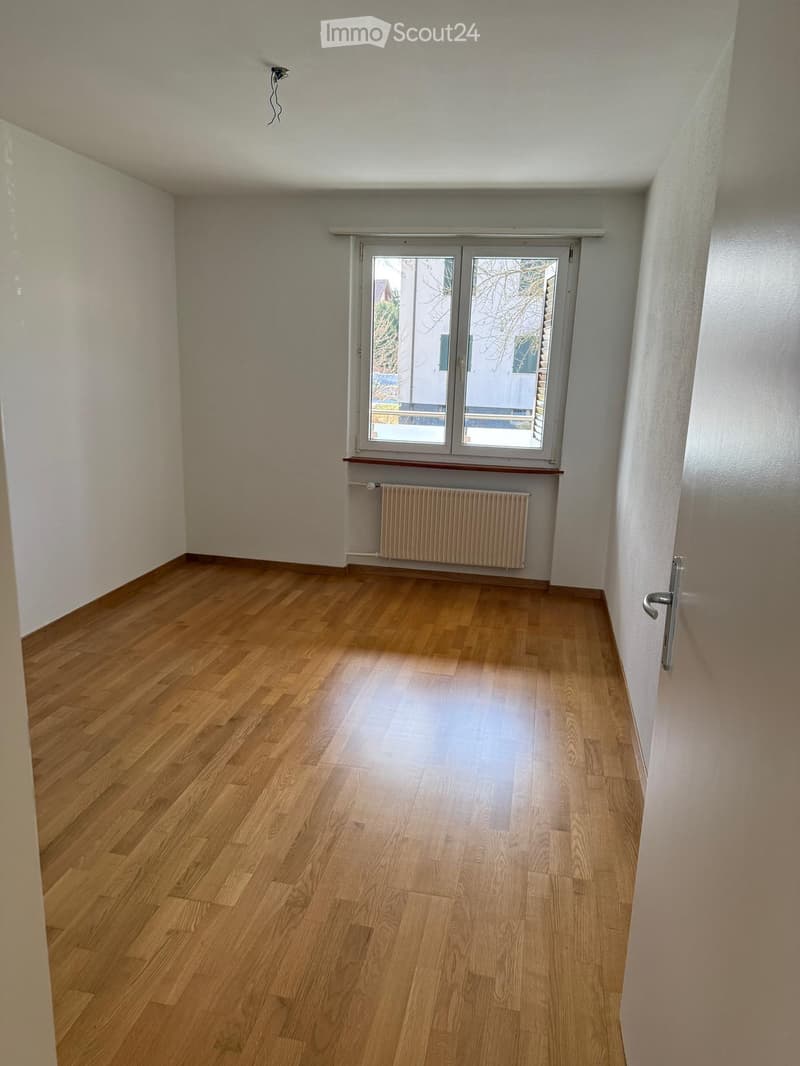 Empty room with wooden flooring, white walls, a window with a radiator below, and a door handle visible on the right side.