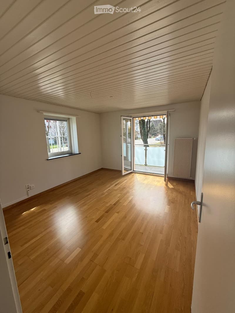 Empty room with wooden flooring, white walls, large window, and sliding glass door leading to a balcony.