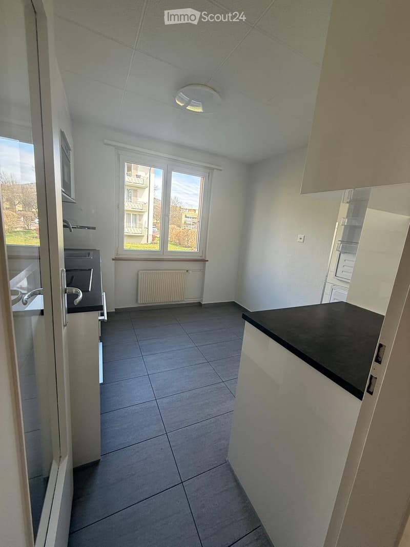 Empty kitchen with tiled floor, white walls, and a window overlooking a balcony. Basic cabinetry and countertop are present, but no appliances or furniture.