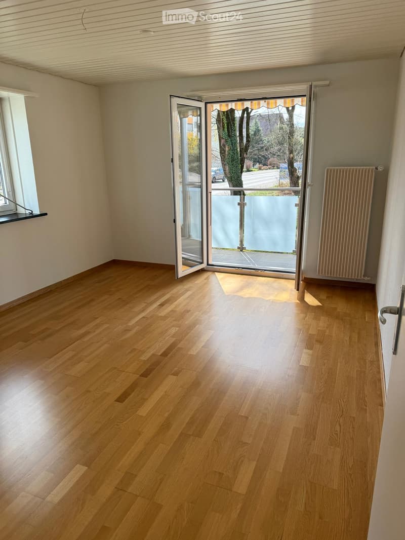 Empty room with light wooden flooring, white walls, large sliding glass door leading to a balcony, and a radiator on the right side.