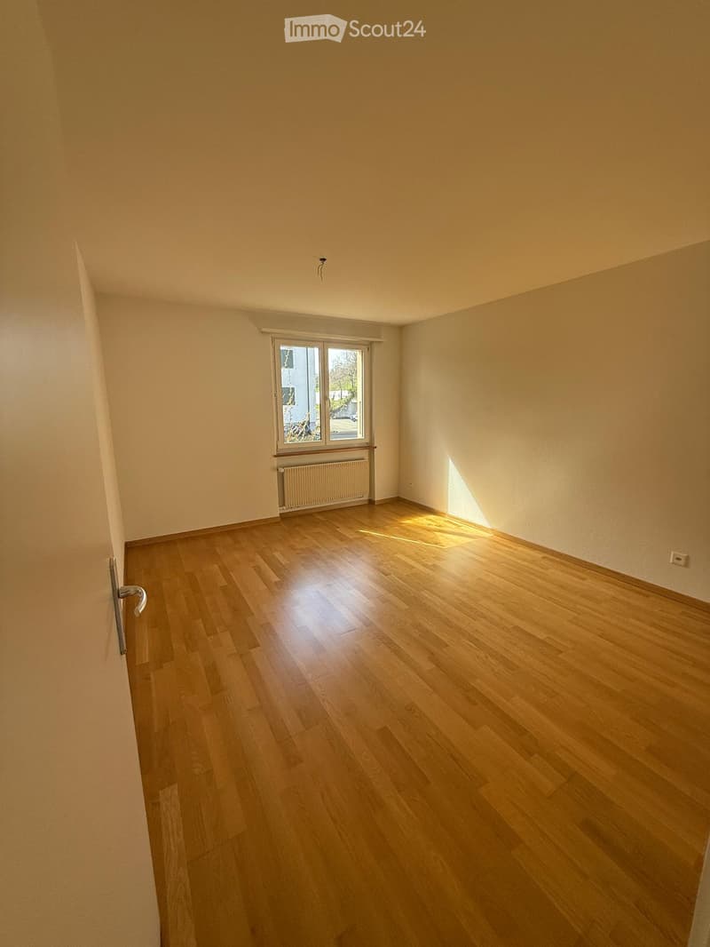 Empty room with wooden flooring, white walls, and a window with a radiator below it.