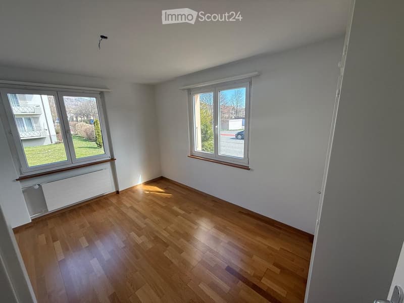 Empty room with wooden flooring, white walls, and large windows offering a view of the outside area.