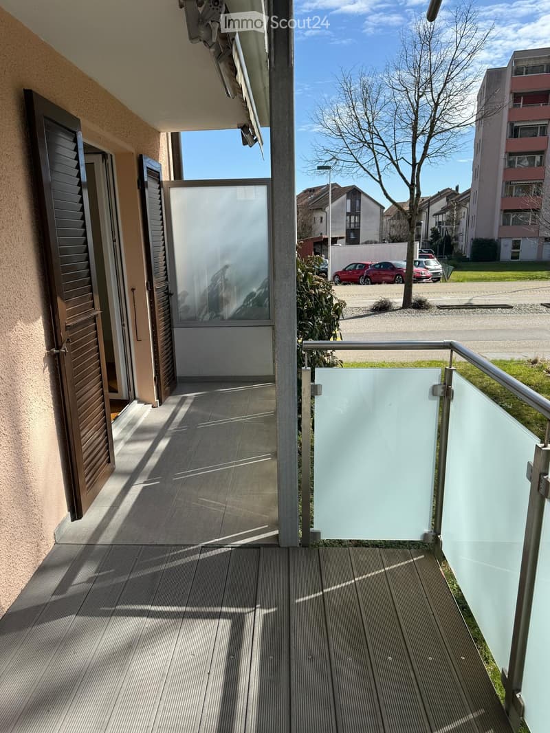 Balcony with wooden decking, glass railing, and open view of the street and surrounding buildings.