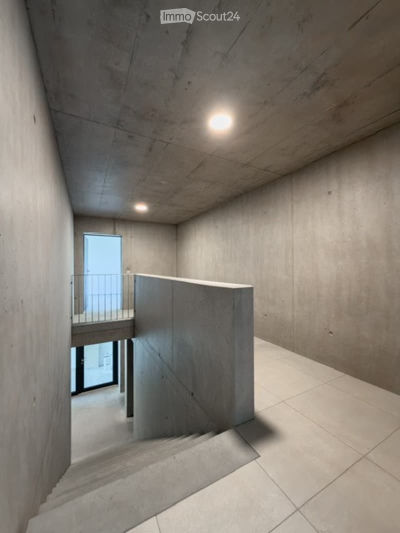 Minimalist concrete hallway with a staircase leading upwards, featuring recessed lighting and a small balcony area visible in the background.