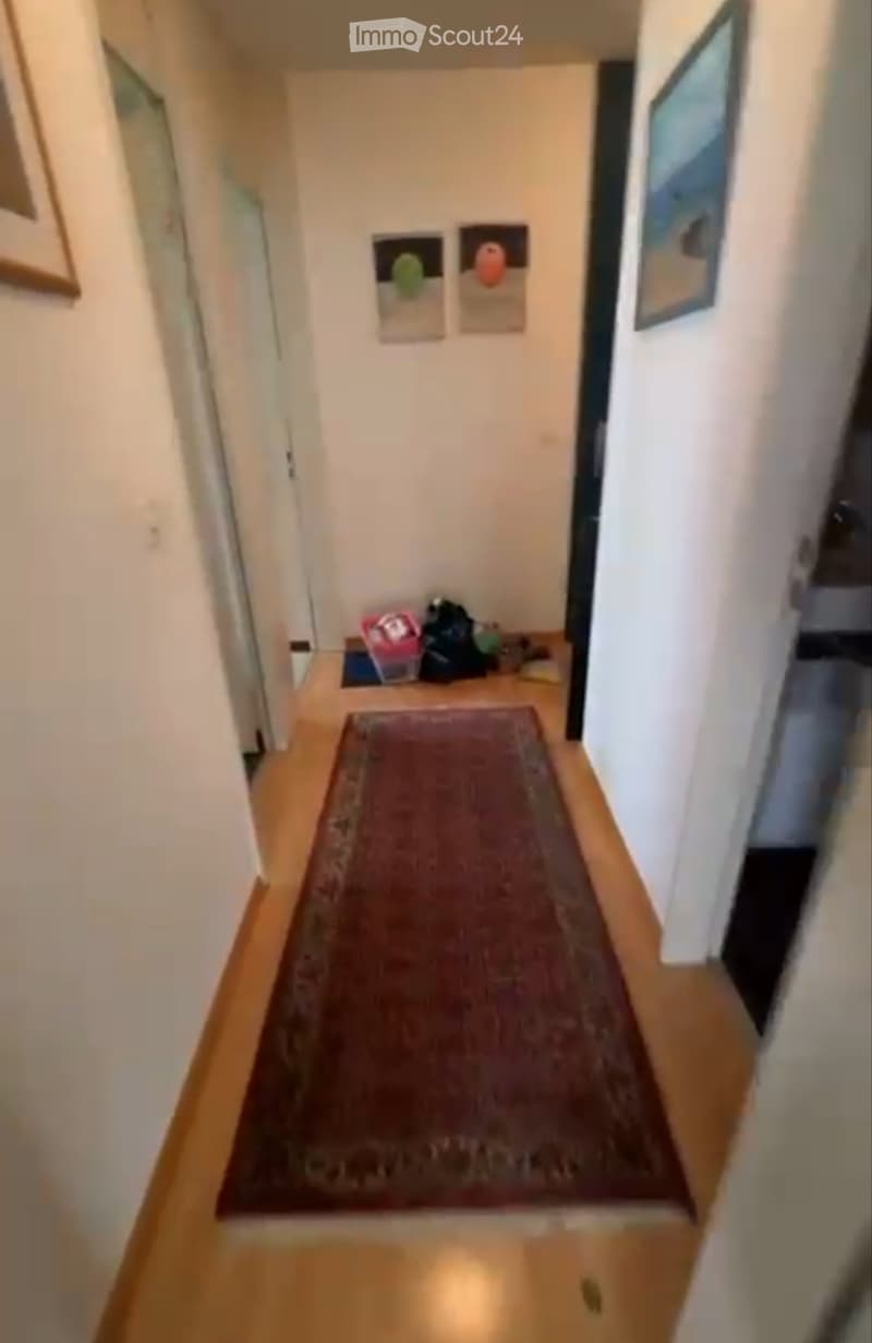 A narrow hallway with wooden flooring, a red runner rug, and several doors on either side. The walls are painted white and feature framed artwork and small decorative items.