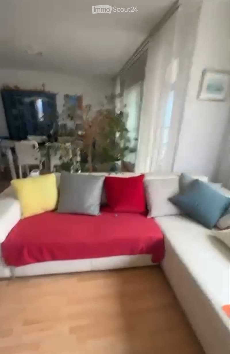 A cozy living room with a white sofa covered in a red blanket and colorful pillows, wooden flooring, and a view of another room through an open doorway.
