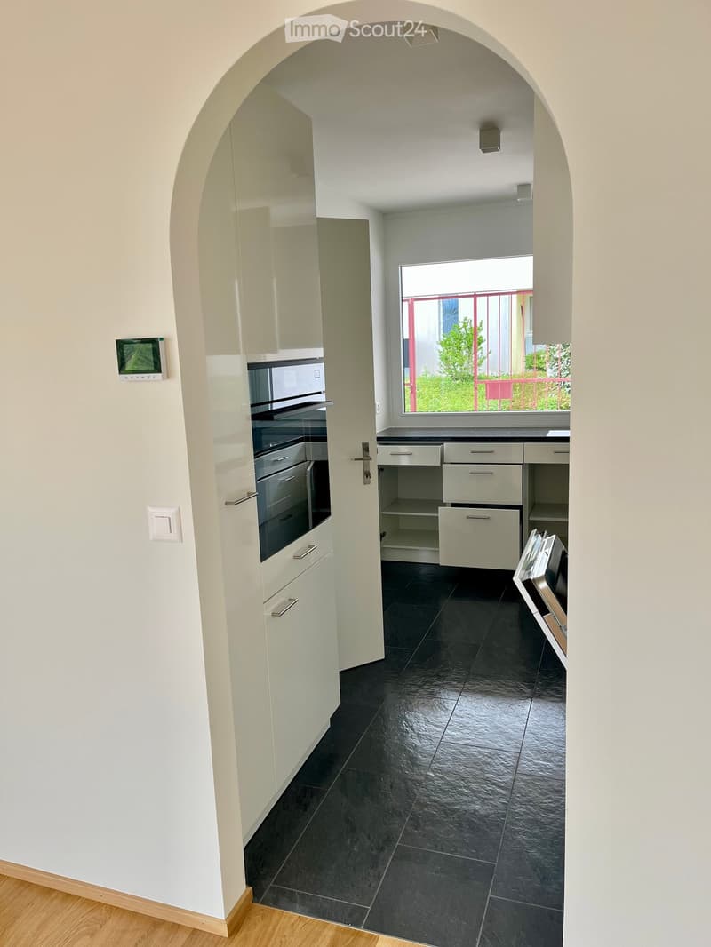 Modern kitchen with white cabinets, black tiled floor, and built-in oven. Features an arched doorway leading to a balcony with a view of greenery.