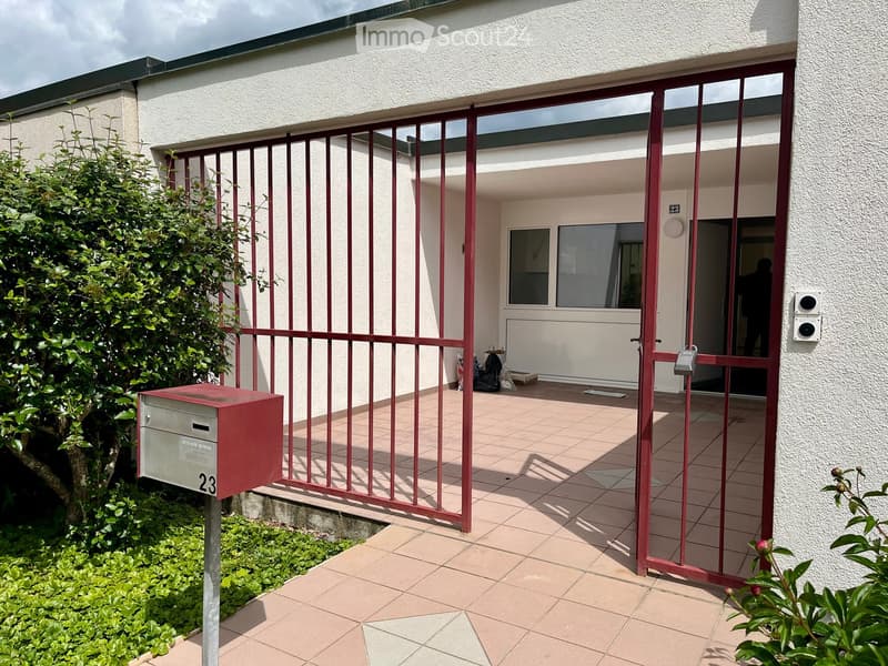Entrance area of a building with a red metal gate, tiled floor, mailbox, and greenery on either side.