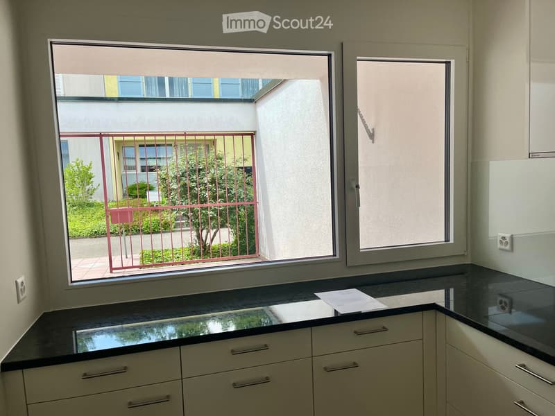 Modern kitchen with white cabinets, black countertop, and large windows offering a view of a garden area.