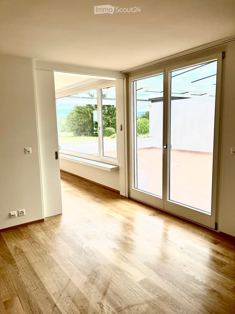 Empty room with light wooden flooring, large sliding glass doors leading to a balcony, and a view of greenery and the sea.