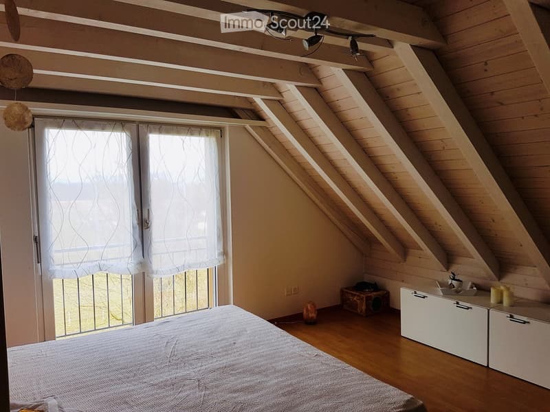 Cozy attic bedroom with wooden slanted ceilings, large window with white curtains, bed, nightstand, and decorative items.