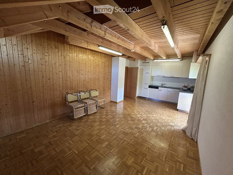 A room with wooden walls and a wooden floor, featuring two chairs, a small stool, a kitchen area with cabinets, a refrigerator, a sink, a faucet, and a stove.