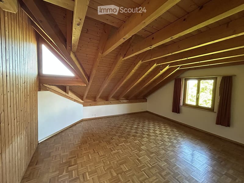 Empty attic room with wooden beams, herringbone wooden flooring, and large windows with curtains.