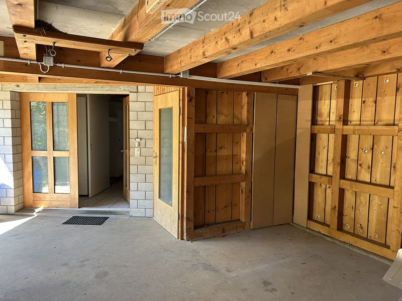 Empty room with exposed wooden beams, concrete floor, and wooden doors leading to another space.