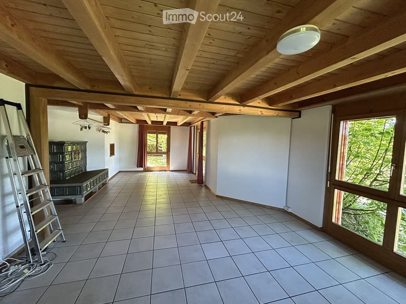 Empty room with exposed wooden beams on the ceiling, tiled floor, large windows, and a ladder leaning against the wall.