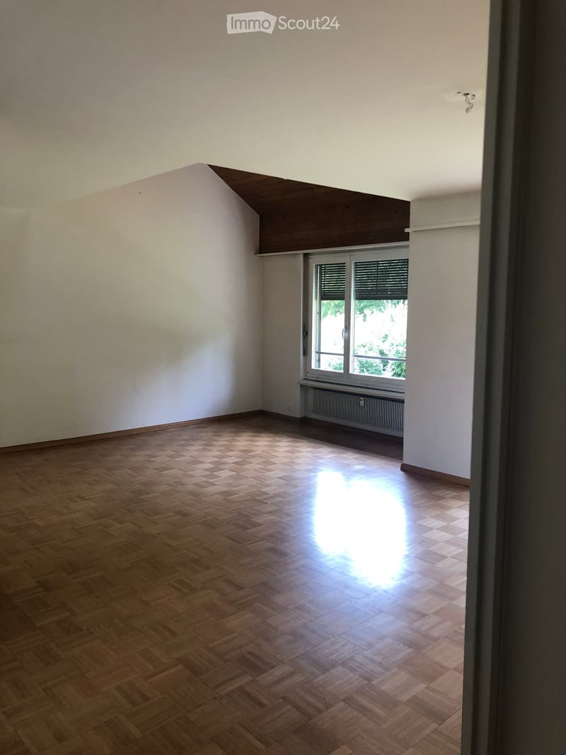 Empty room with wooden herringbone flooring, white walls, and a window with blinds. Natural light streams in, illuminating the space.