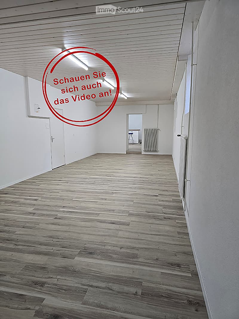 Empty hallway with wooden flooring and white walls, featuring a visible door and a small room at the end with a radiator and a chair.