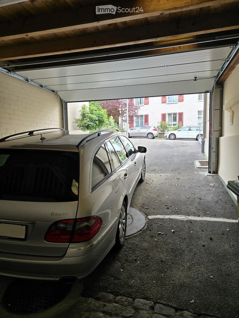 Garage with an open automatic door, a parked car inside, and a view of the street with other parked cars and residential buildings.