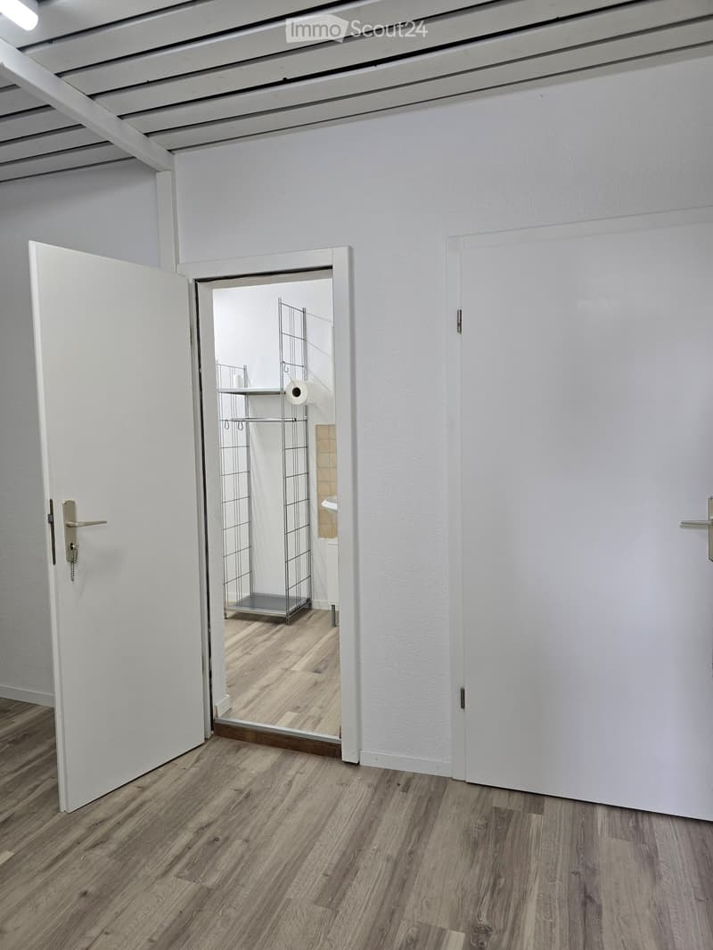 Empty hallway with white walls, wooden flooring, and an open door leading to a small, unfurnished room with a metal shelving unit and a toilet roll holder.