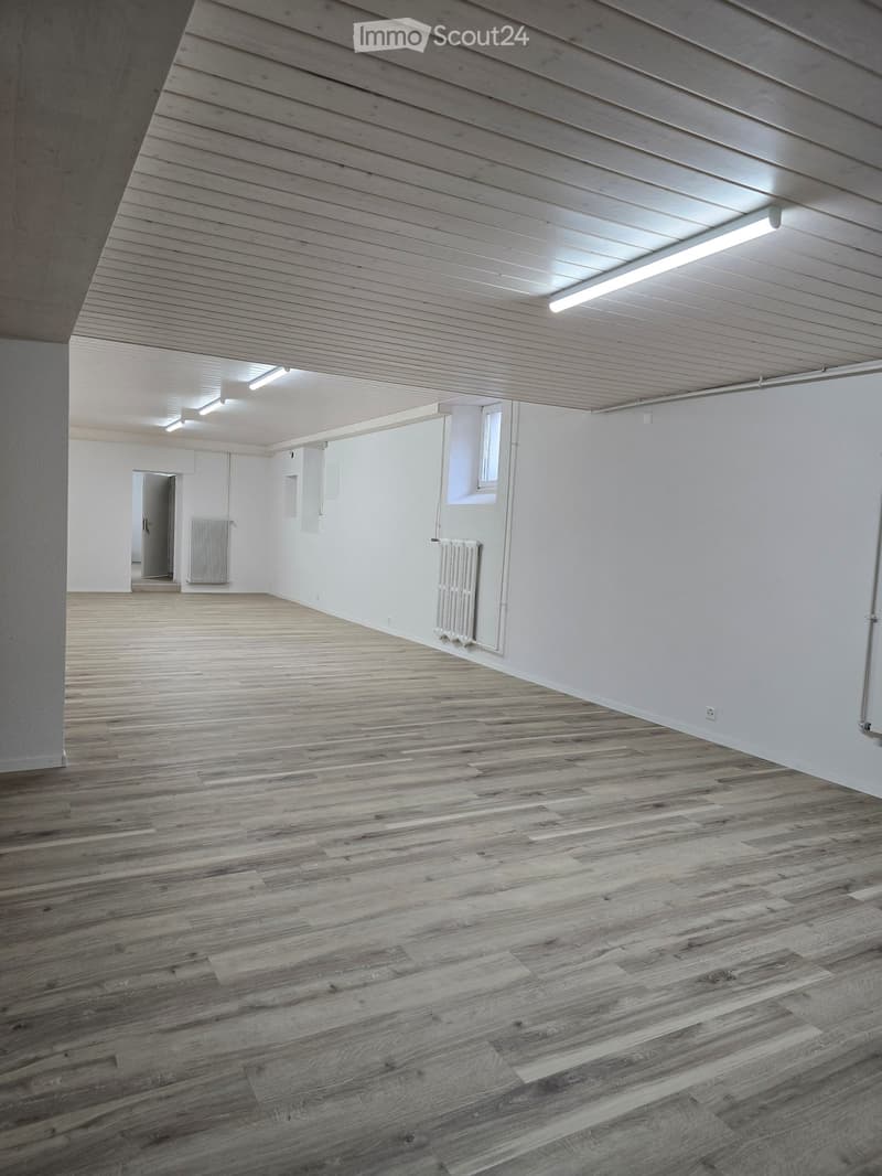 Empty room with wooden flooring, white walls, and a slanted wooden ceiling. Features include a radiator, a small window, and a doorway leading to another space.