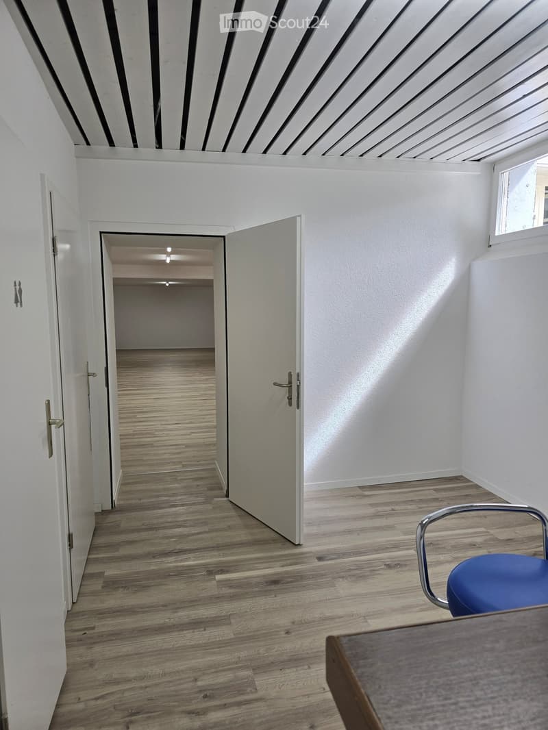 A modern, minimalist hallway with light wooden flooring, white walls, and a slanted ceiling with black and white geometric patterns. A blue chair and a wooden table are visible in the foreground, with an open door leading to another room.