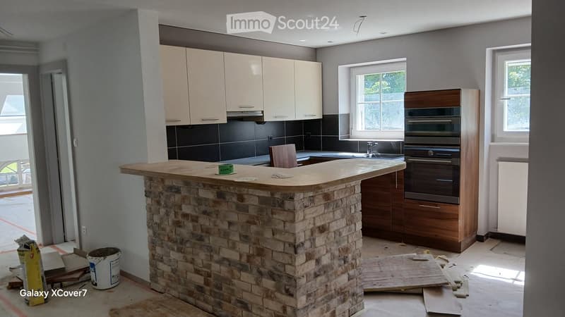 Modern kitchen with white cabinets, a stone island, and a built-in oven. The space features a sink with a faucet, a wooden stool, and a partially constructed island with a stone base.
