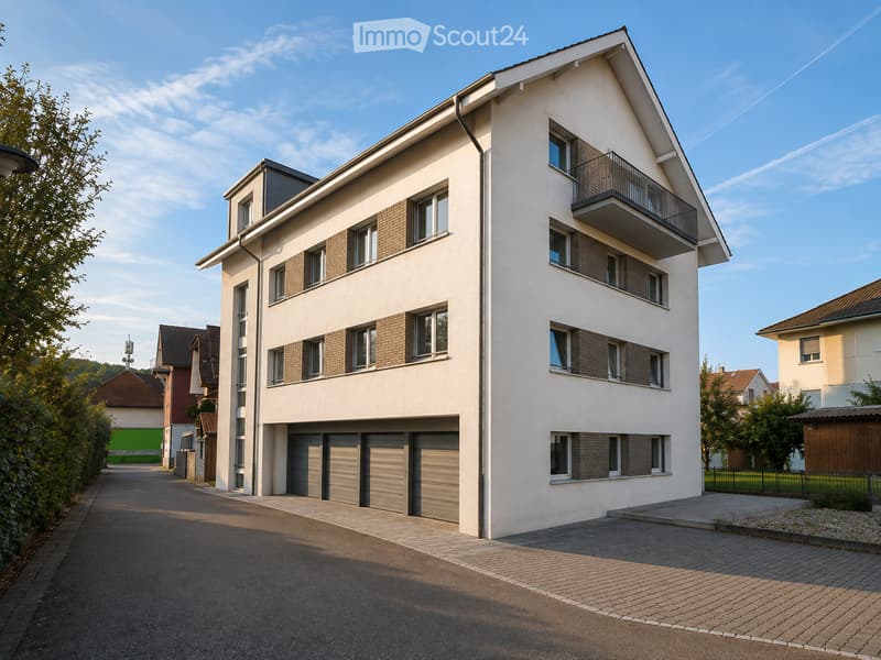 Modern two-story residential building with white facade, multiple windows, and a balcony on the upper floor. Features a garage with three doors and is situated in a quiet neighborhood with paved driveway and surrounding greenery.